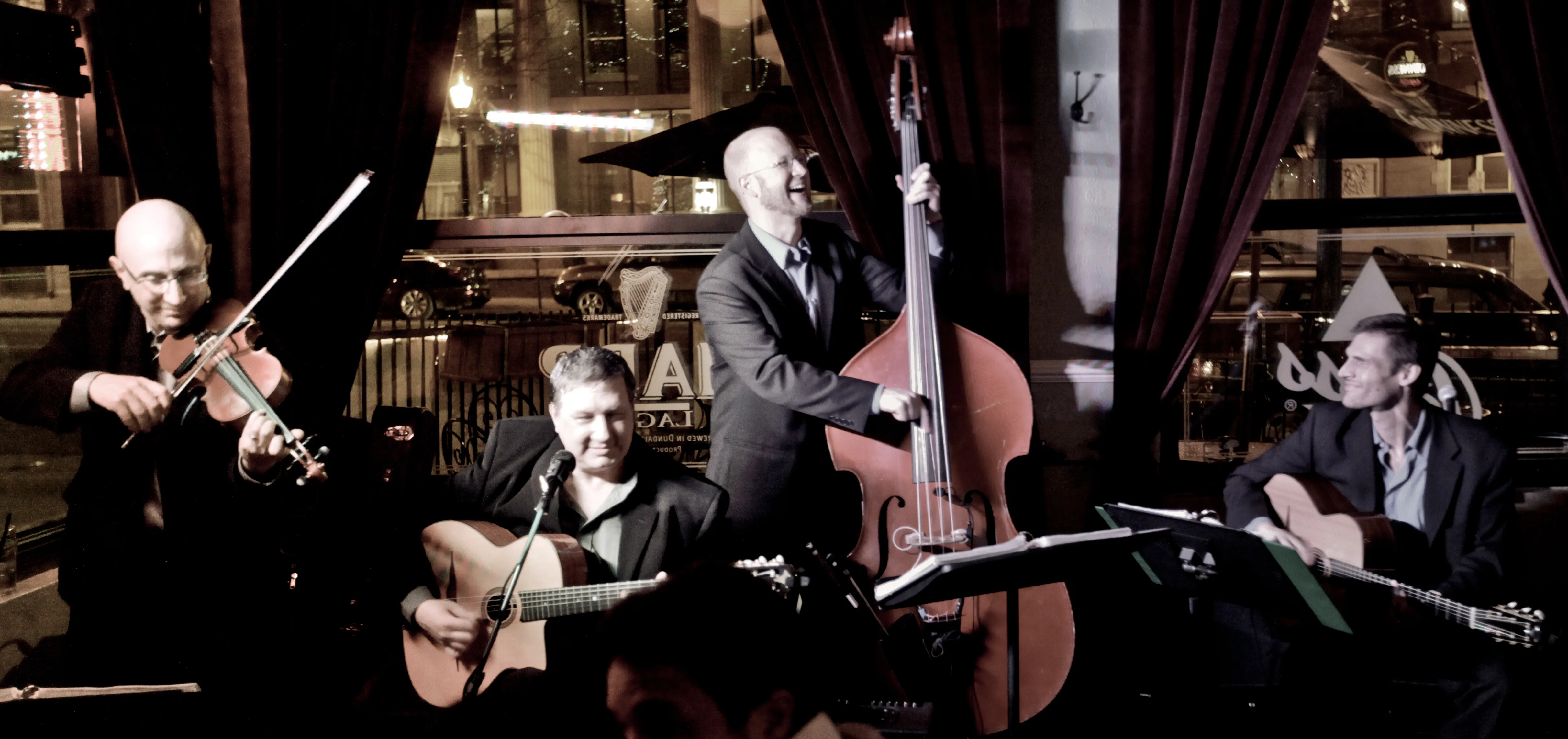 The band Gypsy Swing Revue performing live against a city backdrop, with violin, lead guitar, upright bass, and rhythm guitar.